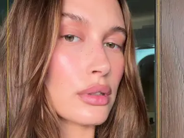 Zbliżenie na twarz Hailey Bieber z delikatnym makijażem i piegami. Jej skóra lśni, a usta są pełne.