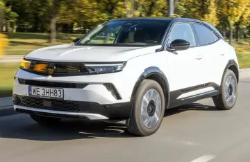 Biały Opel Mokka 2023 z czarnym dachem pędzi po drodze, otoczony jesiennymi drzewami.