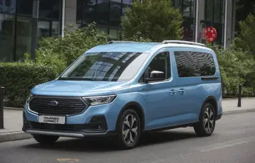 Niebieski Ford Tourneo Connect zaparkowany na ulicy, z budynkiem i zielenią w tle.