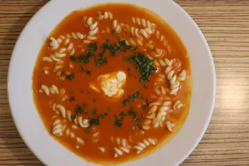 Pomidorowa z makaronem fusilli, udekorowana śmietaną i natką pietruszki, na białym talerzu.