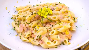 Pasta penne cremosa con trozos de salmón rosado, ralladura de limón y flores amarillas.