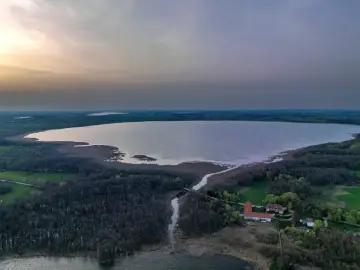 Rozległe jezioro Tuchlin otoczone lasem, z widocznym budynkiem i drogą. Zachód słońca maluje niebo.