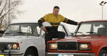 Mężczyzna w kombinezonie rajdowym opiera się o czerwonego Łada 2107, obok stoi biały Fiat 125p.