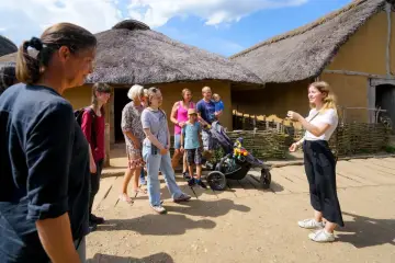 Familienurlaub in Haithabu: Eine Gruppe erkundet historische Häuser mit Strohdächern, während eine junge Frau eine Führung gibt.