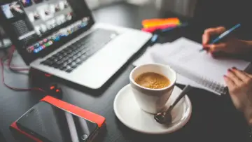 Tazza di caffè, laptop, smartphone e mani che scrivono su un quaderno. Questo è cosa significa da remoto: lavorare ovunque.
