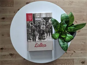 Okładka "Lalki" Bolesława Prusa z ilustracją przedstawiającą scenę z XIX wieku, gdzie widać grupę ludzi w eleganckich strojach. W tle sklep "Riegert". Obok książki stoi zielona roślina.