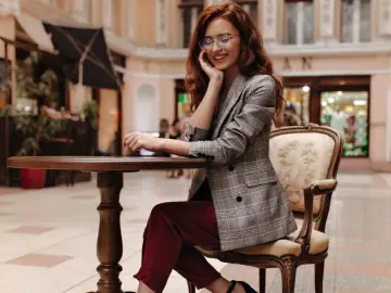 Elegancka kobieta w okularach i marynarce w kratę, prezentująca stylizacje z bordowymi spodniami, siedzi przy stoliku w kawiarni.