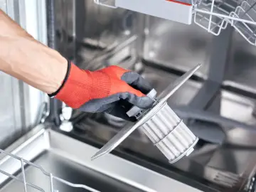 Ręka w rękawicy wyjmuje filtr zmywarki. Regularne czyszczenie filtra to klucz do świeżości naczyń, dlatego odświeżacze do zmywarek są zbędne jeśli dbasz o czystość filtra.