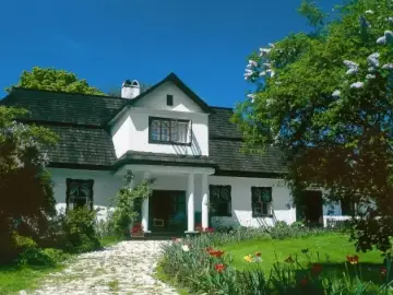 Biały dom z gontowym dachem i zabudowanym gankiem, otoczony kwitnącym ogrodem i drzewami.