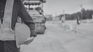 Budowlaniec w kamizelce odblaskowej trzyma kask. W tle walec drogowy i teren budowy, być może przygotowania do wywłaszczenia.