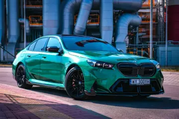 Błyszczące, zielone BMW M5 G90 z czarnymi felgami stoi na tle przemysłowych rur.