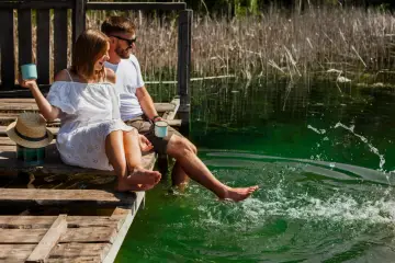 Para na drewnianym pomoście nad jeziorem Zagnanie, delektując się chwilą. Kobieta w białej sukience i mężczyzna w okularach przeciwsłonecznych zanurzają stopy w orzeźwiającej wodzie.