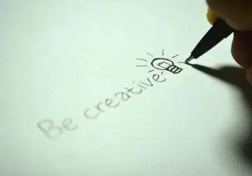 Dłoń pisze długopisem "Be creative" i rysuje żarówkę, symbolizującą pomysł. To inspiracja w stylu popularnonaukowym.