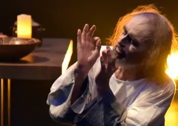Aktor w białej szacie, z rozwianymi włosami, gestykuluje. Scena przypomina "Dziady" w reżyserii Rollisona.