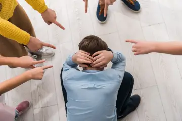 Chłopiec skulony na podłodze, otoczony przez palce wskazujące na niego. To obraz **bullyingu**, który sprawia, że czuje się samotny i przygnębiony.