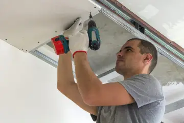 Pracownik w białych rękawiczkach wykonuje montaż płyt gipsowych na wkręty do metalowego stelaża sufitu podwieszanego.