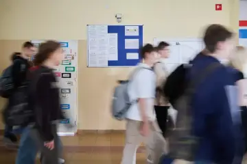 Rozmazani uczniowie idący szkolnym korytarzem obok tablic ogłoszeń.