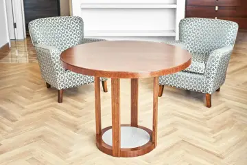Okrągły stolik z forniru z klejem, otoczony dwoma fotelami w geometryczny wzór, stoi na parkiecie w jodełkę.
