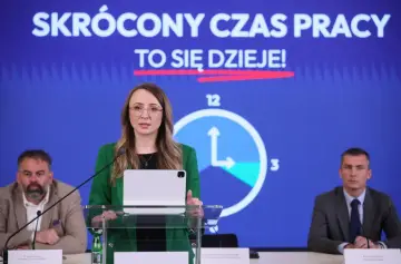 Kobieta przemawia na tle hasła "SKRÓCONY CZAS PRACY TO SIĘ DZIEJE!". Obok niej zegar symbolizujący skrócenie czasu pracy.