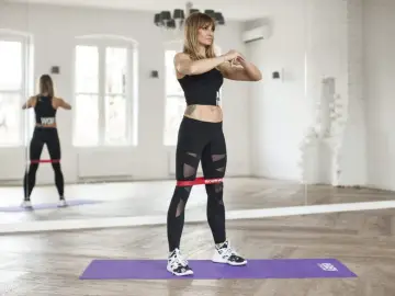 Kobieta wykonuje ćwiczenia z gumami mini band, wzmacniając nogi. Widoczna jest jej sylwetka w lustrzanym odbiciu.