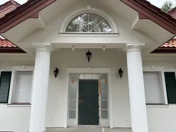 Elegancki pilaster zdobi fasadę domu, wspierając dach nad wejściem. Obok okna i drzwi, tworzy klasyczną kompozycję.