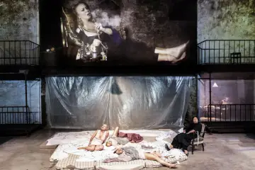 Na scenie teatralnej, w tle projekcja kobiety z fajką, a na dole leżące kobiety. Aluzja do przeszłości.