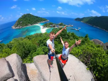 Dwóch mężczyzn robi selfie na tle tropikalnej wyspy z turkusową wodą i plażą.