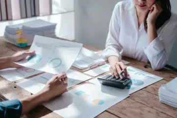 Dwie osoby analizują wykresy i raporty, jedna korzysta z kalkulatora. Rozważają amortyzację jednorazową, planując finanse.
