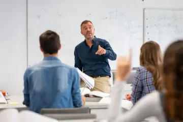 Nauczyciel matematyki tłumaczy zadania z geometrii uczniom w 3 klasie liceum. Na tablicy widać wzory i rysunki.