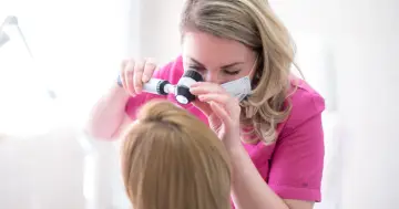 Trycholog w różowej bluzce bada skórę głowy pacjentki dermatoskopem.