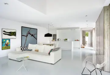 Nowoczesny salon z białym narożnikiem i minimalistyczną kuchnią. Betonowe ściany z żywicy epoksydowej dodają industrialnego charakteru.