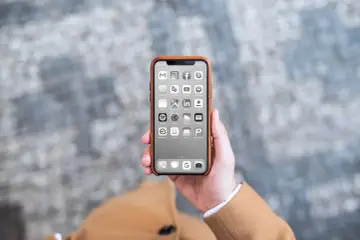 Dłoń trzyma iPhone'a z czarno-białym ekranem i ikonami aplikacji.