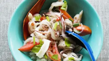 Ensalada tailandesa con fideos de cristal, calamares, tomate y cebolla, servida en un cuenco azul. El plato de sepia en salsa se ve fresco y apetitoso.