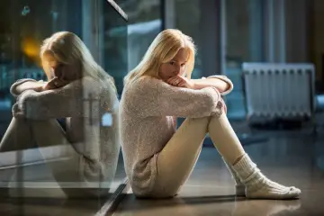 Blondynka siedzi przy oknie, pogrążona w myślach. Jej odbicie w szybie podkreśla uczucie samotności.