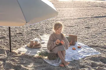Dziewczynka na plaży pod parasolem, jedząca owoce. Idealny przykład, co zabrać na wakacje nad morze z dzieckiem: koc, owoce, ananas i arbuz.