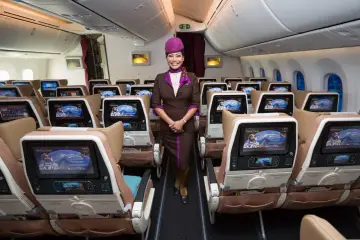 Uśmiechnięta stewardessa Etihad Airways w brązowym mundurku stoi w rzędzie foteli. Ekraniki pokazują astronautę. Dobre etihad airways opinie.