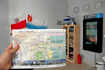 Ręka trzyma mapę "Gdynia w jeden dzień", pokazującą trasę spaceru po Gdyni. W tle flagi i zegary.
