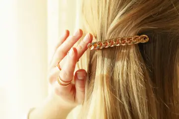 Złota klamra w kształcie łańcuszka ozdabia blond włosy. Dłoń z pierścionkami delikatnie je układa, pokazując, jak upiąć włosy klamrą.