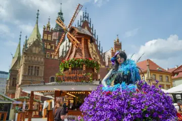 Barwna postać w kwiecistej sukni na jarmarku świętojańskim we Wrocławiu, z wiatrakiem w tle.