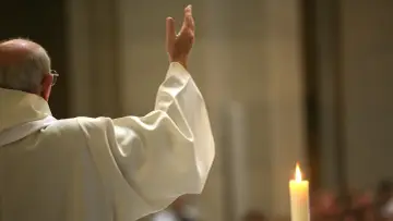 Kapłan w białej szacie podnosi rękę. Czy opuszczając jedną mszę można iść do komunii? Płonąca świeca obok.