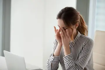 Kobieta z zapaleniem spojówek masuje oczy, zmęczona pracą przy komputerze.