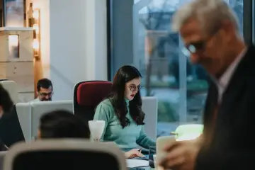 Kobieta w okularach pracuje przy biurku, obok mężczyzna w garniturze.