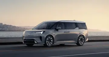 Szary Volvo EM90 jedzie wzdłuż wybrzeża o zachodzie słońca.