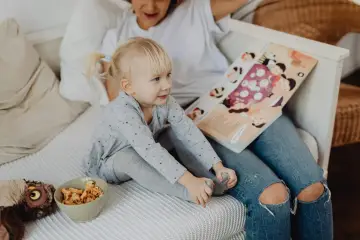 Mała dziewczynka z blond kucykami słucha bajki czytanej przez opiekunkę. Zastanawiasz się, ile zarabia opiekunka dla dziecka?