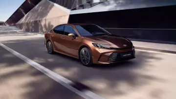 Brązowa Toyota Camry 2.5 Hybrid sunie po drodze, otoczona nowoczesną architekturą i cieniem drzew.