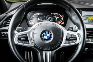 Kierownica BMW z cyfrowym kokpitem i przyciskami tempomatu. HBA (asystent świateł drogowych) włączony, widoczny na ekranie.