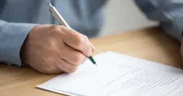 Dłoń podpisuje umowę dzierżawy. Dokument leży na drewnianym biurku, a osoba używa długopisu.