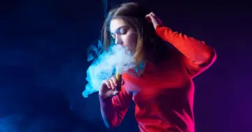 Joven exhala humo de un vaper, rodeada de luces azules y moradas.