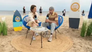 Wywiad na plaży w Świnoujściu. Dziennikarka z mikrofonem rozmawia z mężczyzną siedzącym na krześle. W tle widać morze i spacerujących ludzi.
