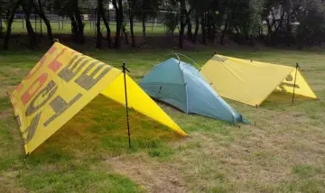 Żółty tarpa z napisem "OUTDOOR" i zielony namiot na trawie.